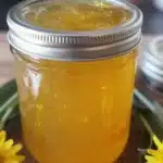 A jar of homemade dandelion jelly on a wooden table with dandelion flowers.