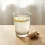 Finished natural GLP-1 drink in a clear glass with chia seeds visible, a thin lemon slice on the rim, and a small piece of fresh ginger root alongside on a wooden surface in morning light