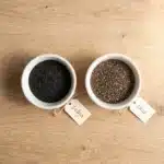 Side-by-side comparison of sabja seeds and chia seeds in two small white bowls on a wooden surface, showing their similar appearance before soaking